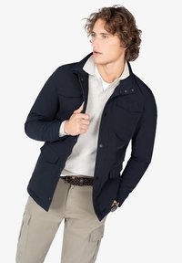 Chaqueta azul marino con cierre de cremallera y botones a presión, que cuenta con dos bolsillos en el pecho y un diseño elegante, llevada sobre un suéter ligero.