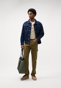 Jeansjakke med knaplukning foran, lysebeige t-shirt, olivengrønne bukser og brune sko. Har en kviltet grøn tote bag.