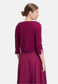 Gilet fuchsia court à manches trois-quarts, avec fentes sur les côtés. Tissu doux et texturé qui complète la robe fuchsia fluide.