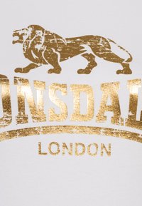 White t-shirt featuring a gold foil lion graphic and bold text reading "Lonsdale" and "London," with a distressed texture for visual interest.