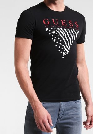 Czarny dopasowany t-shirt z czerwonym napisem "GUESS" oraz białym trójkątnym wzorem zawierającym gwiazdy i paski, noszony z niebieskimi dżinsami.