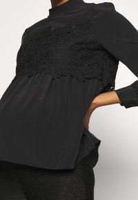 Czarna bluzka z teksturową koronkową wstawką na wysokości klatki piersiowej, z długimi rękawami i wysokim dekoltem. Miękka, płynąca tkanina poniżej talii.