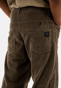 Pantalones de pana marrón con una textura acanalada, que cuentan con dos bolsillos traseros y un pequeño parche con el logotipo. La tela es suave y duradera.