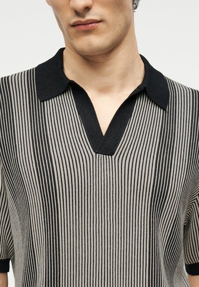 Homme portant une chemise en tricot à manches courtes avec des rayures verticales noires et grises et un col noir, debout devant un fond uni.