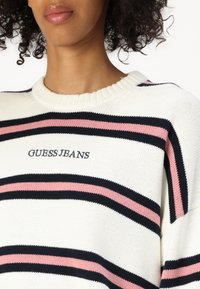 Valge kudum, millel on horisontaalsed triibud tumesinisest, roosast ja keskel "GUESS JEANS" logo. Ribitud kaelus.