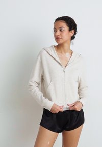 Casaco de capuz em bege claro com fecho, capuz e bolsos frontais, feito de um tecido suave. Combinado com calções de satin pretos para um visual descontraído.