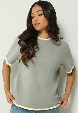 Femme aux longs cheveux foncés portant un t-shirt gris clair avec une bordure jaune pâle aux manches et à l'encolure, debout devant un fond uni.