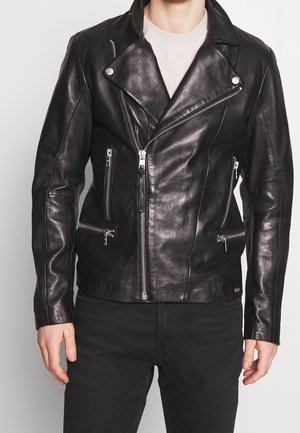 Czarna skórzana kurtka motocyklowa z asymetrycznym zamkiem błyskawicznym, cztery kieszenie na zamek, noszona na beżowej bluzce i zestawiona z czarnymi spodniami.