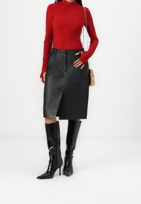 Rotes langärmliges Oberteil, figurbetont; schwarze Leder-Midi-Rock mit Schlitz vorne; schwarze glänzende kniehohe Stiefel mit Absatz; goldene Handtasche.