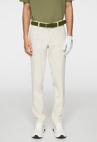 Pantaloni beige con una superficie liscia, indossati con una cintura intrecciata verde. Abbinati a sneakers bianche e a un guanto bianco, con logo.