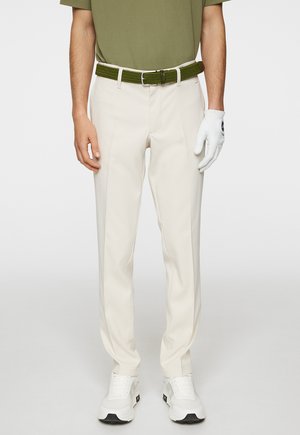 Beige trousers with a smooth texture, worn with a green woven belt. Paired with white sneakers and a white glove, featuring a logo.