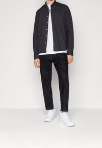 Camicia nera con bottoni dorati, abbinata a jeans neri testurizzati e sneakers bianche, posizionata su uno sfondo neutro.