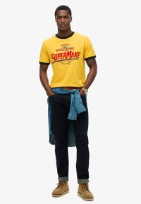 Camiseta gráfica amarilla con ribetes negros, camisa de mezclilla azul atada a la cintura, jeans oscuros y botas de color marrón claro. Presenta texto en rojo brillante en la camiseta.