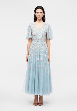 Kobieta w jasnoniebieskiej, midi sukience z haftem w kwiaty, z dekoltem w serek, krótkimi falującymi rękawami i przezroczystym materiałem, stojąca na jednolitym tle.