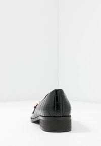 Mocassini neri con texture di coccodrillo, tacco basso e dettagli in hardware dorato. Materiale liscio e lucido con una forma strutturata.