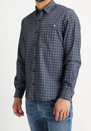 Mężczyzna ubrany w ciemnoszarą i czarną koszulę w kratę z długim rękawem oraz niebieskie dżinsy, stojący na tle jednolitego białego tła.