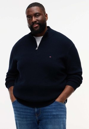 Μαύρος άνδρας με γένια, φοράει ναυτικό μπλε πουλόβερ με φερμουάρ στη μέση και μπλε τζιν, στέκεται με τα χέρια στις τσέπες μπροστά σε λευκό φόντο.