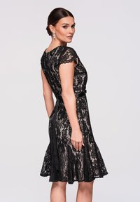 Femme portant une robe noire en dentelle jusqu'aux genoux avec des manches courtes et une ceinture, regardant par-dessus son épaule sur un fond uni.