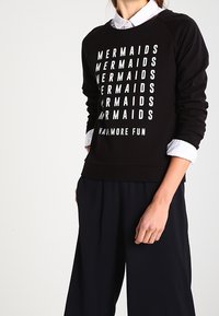 Persona che indossa una felpa nera con il testo bianco che ripete "MERMAIDS" e "HAVE MORE FUN", abbinata a pantaloni neri e una maglietta bianca.