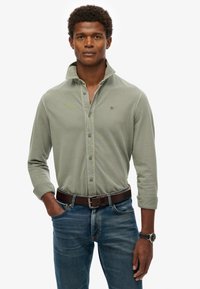 Langärmliges olivgrünes Button-Up-Hemd mit Kragen, kombiniert mit blauen Jeans und einem braunen Ledergürtel, dazu eine Uhr.