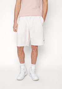 Bílé bavlněné kraťasy se záhyby, obsahující boční kapsy a malou černou Nike logo. Nosič má na sobě bílé tenisky a nenápadnou růžovou košili.