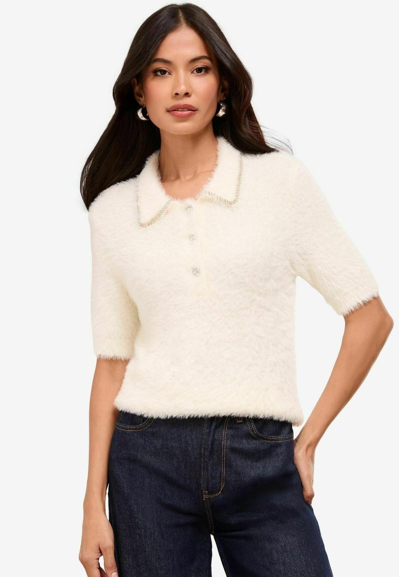 Suéter de polo blanco esponjoso con mangas cortas, botones de perla y un acabado suavemente texturizado. Presenta un cuello con un borde decorativo.