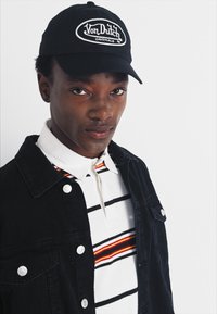Căciulă neagră cu logo-ul "Von Dutch", tricou polo alb și negru cu dungi și accente portocalii, și o jachetă neagră cu nasturi tip snap.
