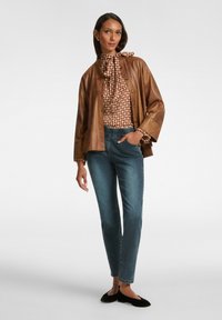 Giacca di pelle marrone con bottoni, blusa a fantasia, jeans blu e scarpe basse nere. La blusa presenta un collo a cravatta e polsini arricciati.
