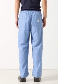 Muž stojící zády, oblečený v modro-bílých tenkých proužkovaných kalhotách s elastickým pasem, zadní kapsou a černých botách.