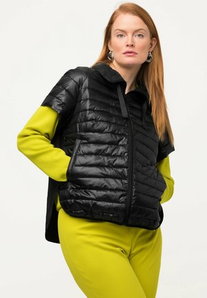 Roodharige vrouw met zilveren oorringen, zwarte gewatteerde jas met korte mouwen over een limoengroene top met lange mouwen en bijpassende broek, handen in de zakken.