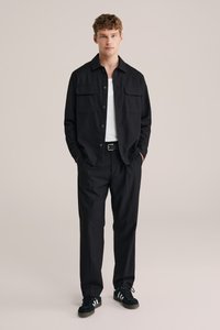 Camicia nera a bottoni con due tasche sul petto, abbinata a pantaloni neri sartoriali e sneakers scure con strisce bianche. Tessuto dalla texture liscia.