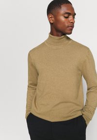 Beige polo sweater i blødt strikmateriale, der har en tætsiddende silhuet og lange ærmer, parret med mørke bukser.