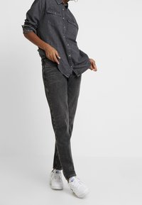Camicia di jeans grigio scuro con due tasche sul petto, bottoni frontali e maniche arrotolate. Abbinata a jeans scuri e sneakers bianche.