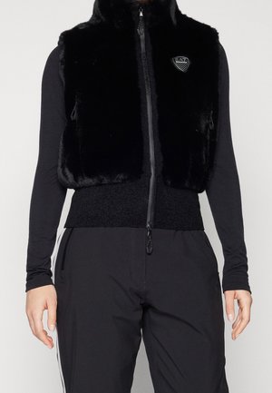 Czarna bezrękawnikowa kamizelka z sztucznego futra z przednim zamkiem błyskawicznym i logo EA7 na klatce piersiowej, noszona na koszuli z długim rękawem oraz czarnych spodniach z bocznymi paskami.