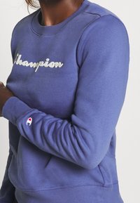 Lila sweatshirt i mjukt tyg med en glansig "Champion"-logotyp i mitten och en liten röd och vit logotyp på ärmen.