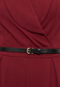 Gros plan sur une robe portefeuille rouge foncé avec une ceinture en cuir noir ornée d'une boucle dorée à la taille.