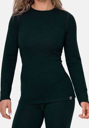 Kobieta ubrana w dopasowaną, ciemnozieloną bluzkę z długim rękawem i pasujące spodnie, widziana od szyi do bioder na jednolitym tle.