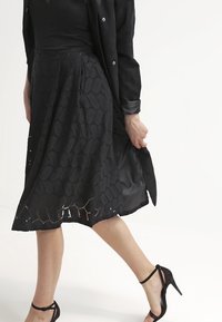 Zwarte knie-lange rok met intricates kantachtige patronen en een geplooid ontwerp, gecombineerd met een aansluitend zwart topje en gespsandalen.