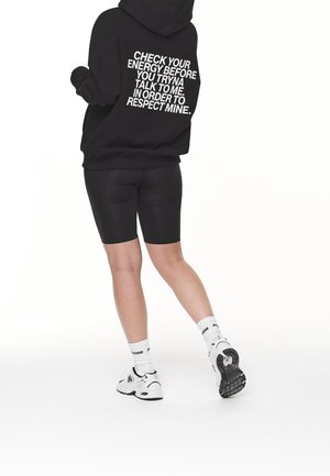 Person iført sort hættetrøje med teksten "Tjek din energi, før du prøver at snakke med mig" går i sorte shorts, hvide sokker og hvide sneakers.