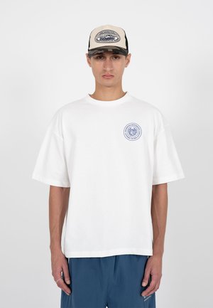 Jonge man met een beige en zwarte pet en een wit oversized T-shirt met een klein rond logo op de borst, staand tegen een effen grijze achtergrond.