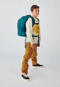 Tarnfarbener Rucksack mit mehreren Taschen und verstellbaren Trägern, getragen von einem Kind in einem kremfarbenen Pullover mit einem gelben Smiley-Gesicht, beigefarbenen Hosen und dunklen Sneakers.