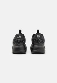 Zapatillas deportivas negras con una parte superior texturizada, costuras en contraste y tiradores, con "SM" bordado en la parte trasera.