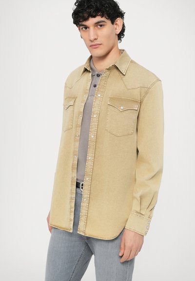 Chemise en denim beige avec fermeture boutonnée, deux poches poitrine, et manches longues avec poignets boutonnés, portée sur un polo gris et un jean gris.
