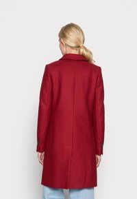Abrigo de lana rojo con cuello con muescas, corte entallado y dos botones en cada puño. Presenta una textura suave y una longitud ligeramente más larga.