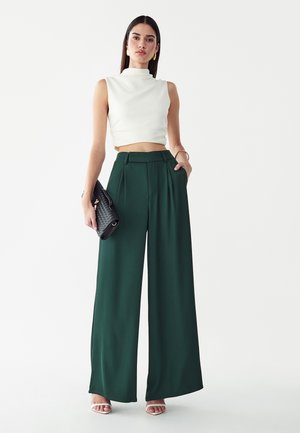 Kvinde iført en hvid ærmeløs crop top, mørkegrønne vidde bukser, hvide sandaler og bærende en sort clutch taske.