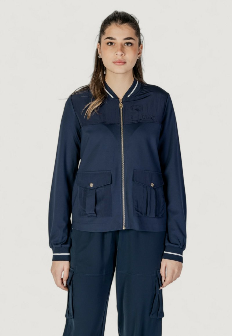 Giacca zip-up navy con colletto a contrasto, tasche frontali e logo ricamato. Presenta una texture liscia e un design aderente.