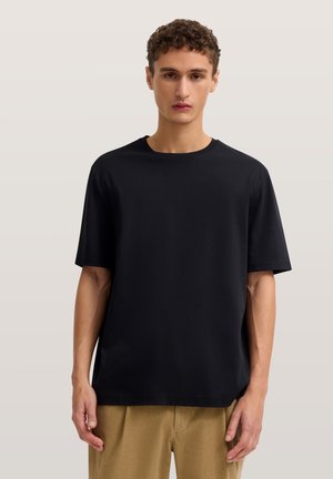 Junger Mann mit kurzen lockigen Haaren steht frontal und trägt ein schlichtes schwarzes T-Shirt mit kurzen Ärmeln und hellbraune Hosen vor neutralem Hintergrund.