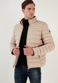 Beige polstret jakke med høj krave, vertikal quiltet design, lynlåslommer og sorte detaljer. Båret over en sort ribbet sweater.