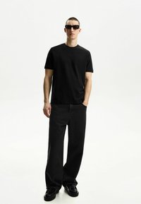 Schwarzes T-Shirt mit kurzen Ärmeln, kombiniert mit locker sitzenden schwarzen Jeans. Modell trägt schwarze Sonnenbrille und schwarze Schuhe. Schlichter Hintergrund.