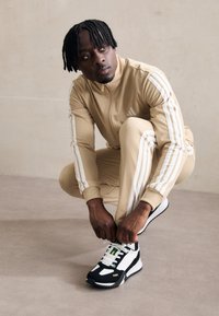 Chándal beige con rayas blancas y cuello con cremallera, combinado con zapatillas en blanco y negro con detalles en verde y dorado.
